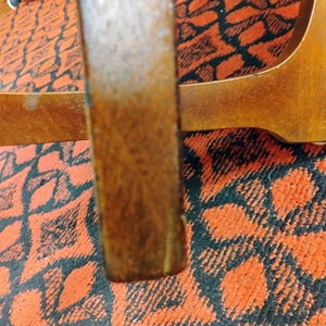 May include: A vertical wooden furniture leg with a medium brown finish and visible wood grain texture. The leg is positioned in front of a horizontal wooden support piece, both set against a vibrant orange and black patterned textile surface.