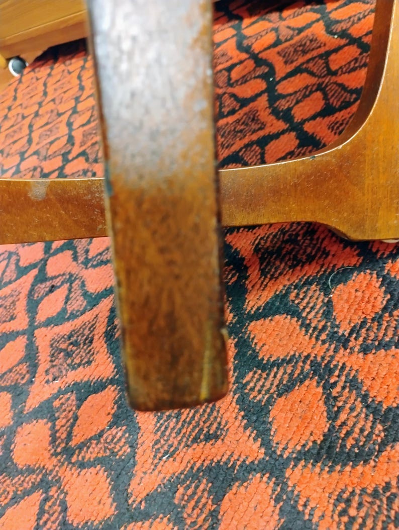 May include: A vertical wooden furniture leg with a medium brown finish and visible wood grain texture. The leg is positioned in front of a horizontal wooden support piece, both set against a vibrant orange and black patterned textile surface.