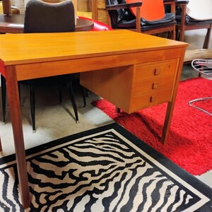 May include: A mid-century modern wooden desk with three drawers. The desk has a rectangular top and tapered legs. The desk is made of light-colored wood and has a warm, inviting tone. The desk is set against a backdrop of other furniture.