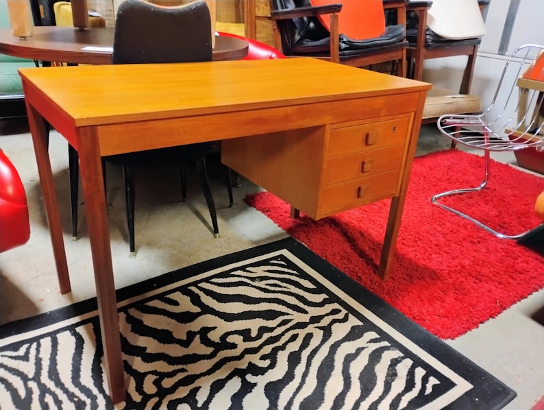 May include: A mid-century modern wooden desk with three drawers. The desk has a rectangular top and tapered legs. The desk is made of light-colored wood and has a warm, inviting tone. The desk is set against a backdrop of other furniture.