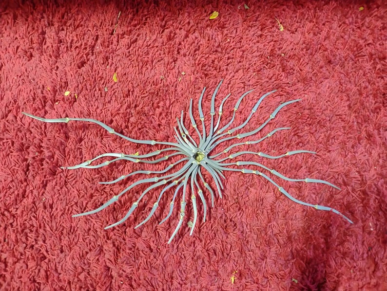 May include: A gray metal decorative piece with a central circular design and radiating, curved arms. The arms have small, yellow accents. The piece is set against a textured, red background.