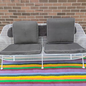 May include: A white wire mesh loveseat featuring a curved frame and a grid pattern. The seating includes two dark gray seat cushions and two matching back cushions, each with white piping along the edges, providing a structured seating area.