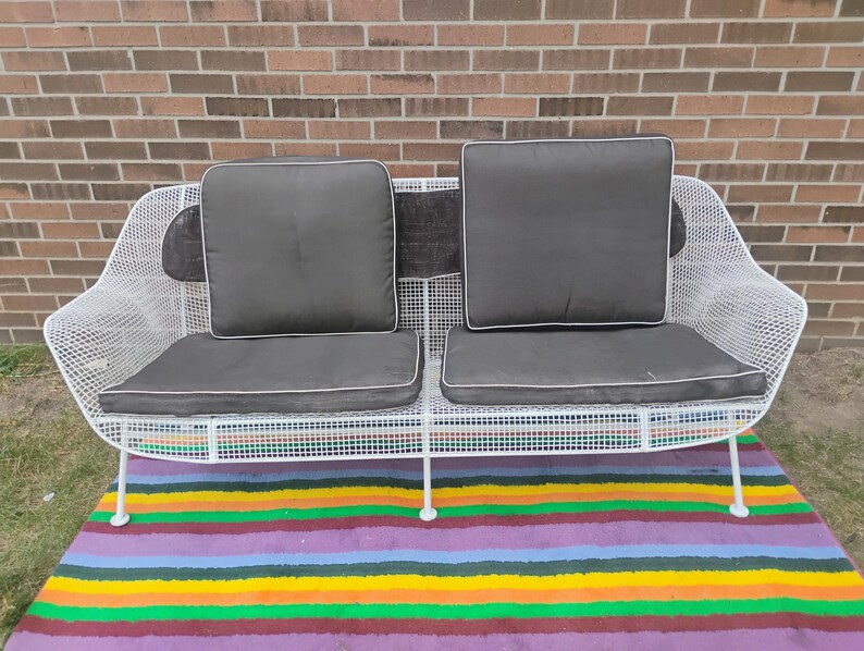 May include: A white wire mesh loveseat featuring a curved frame and a grid pattern. The seating includes two dark gray seat cushions and two matching back cushions, each with white piping along the edges, providing a structured seating area.