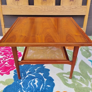 May include: A mid-century modern coffee table with a wood top and a woven wicker shelf. The table has a square top and four legs.