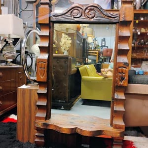 May include: A wooden framed mirror with intricate carvings and a shelf. The mirror is reflecting a room with furniture and a person holding a phone.
