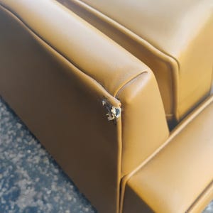 May include: A mustard yellow sofa with smooth upholstery and a visible tear on the top corner edge of the backrest. The couch features clean, straight lines and a structured design with padded sections, showing signs of wear on the corner seam.