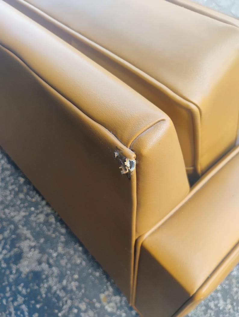 May include: A mustard yellow sofa with smooth upholstery and a visible tear on the top corner edge of the backrest. The couch features clean, straight lines and a structured design with padded sections, showing signs of wear on the corner seam.