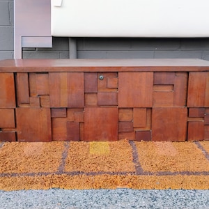 May include: A mid-century modern wooden chest with a geometric design. The chest is made of various sized rectangular blocks of wood, creating a textured surface. The chest is brown and sits on a yellow and brown rug.