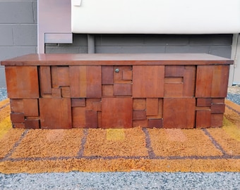 May include: A mid-century modern wooden chest with a geometric design. The chest is made of various sized rectangular blocks of wood, creating a textured surface. The chest is brown and sits on a yellow and brown rug.