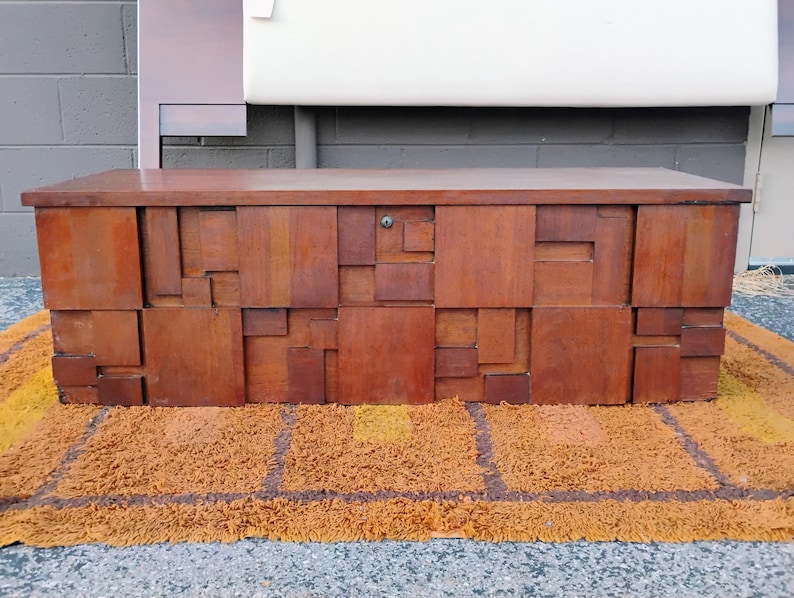 Puede incluir: Un cofre de madera de estilo moderno de mediados de siglo con un dise&ntilde;o geom&eacute;trico. El cofre est&aacute; hecho de bloques rectangulares de madera de varios tama&ntilde;os, creando una superficie texturizada. El cofre es marr&oacute;n y est&aacute; sobre una alfombra amarilla y marr&oacute;n.