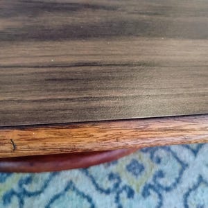 May include: Close-up of a wooden table with a dark brown surface and a lighter brown edge. The table has a visible wood grain pattern. The edge of the table is a lighter shade of brown.