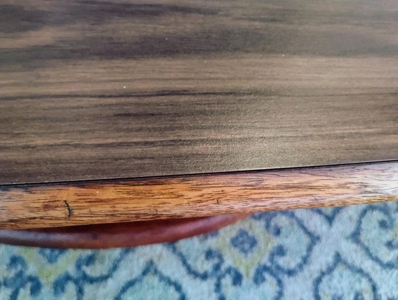 May include: Close-up of a wooden table with a dark brown surface and a lighter brown edge. The table has a visible wood grain pattern. The edge of the table is a lighter shade of brown.