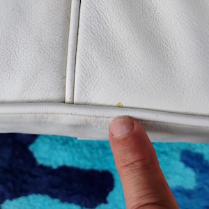 May include: Close-up of a white leather object with visible stitching and a textured surface. A finger is touching the edge of the object. The background features a blue and teal patterned fabric.