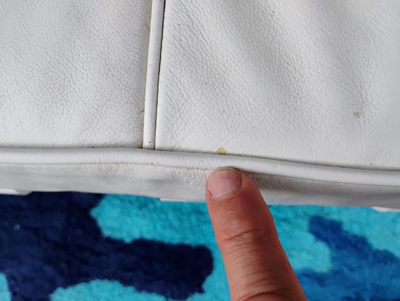 May include: Close-up of a white leather object with visible stitching and a textured surface. A finger is touching the edge of the object. The background features a blue and teal patterned fabric.