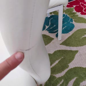 May include: Close-up of a white chair with a curved back and a white metal frame. The chair is on a floral patterned rug with green, red, and blue accents. The chair's surface appears to be smooth.