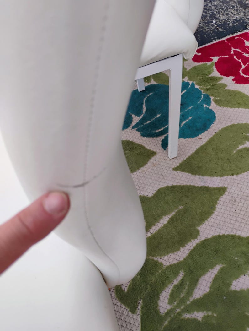 May include: Close-up of a white chair with a curved back and a white metal frame. The chair is on a floral patterned rug with green, red, and blue accents. The chair's surface appears to be smooth.