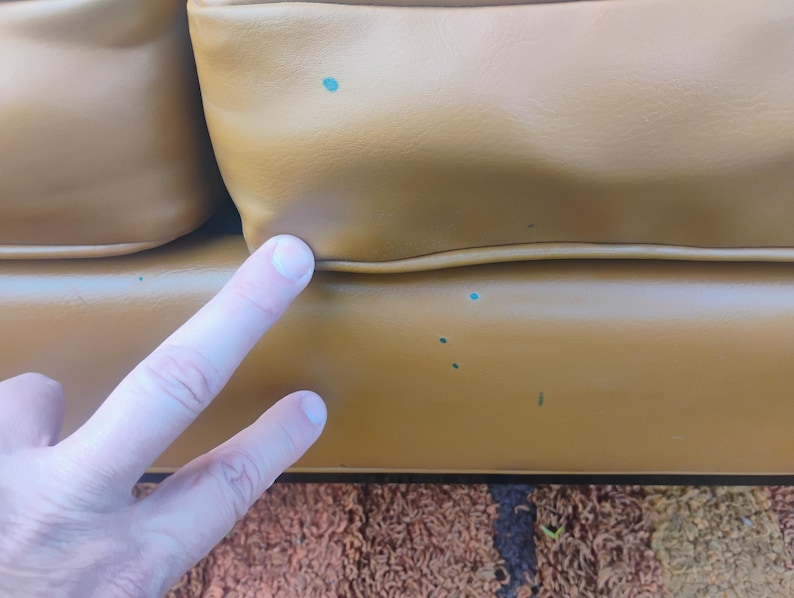 May include: A mustard yellow faux leather sofa cushion featuring several small, distinct teal-colored ink or paint spots on the surface. The material has a smooth texture with visible seams along the edges of the upholstered furniture piece.