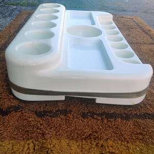 May include: A white plastic organizer tray featuring multiple circular indentations of varying sizes, two rectangular recessed compartments, and one central circular basin, all integrated into a single molded unit with a grey band around the base.