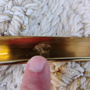 May include: A close-up of a gold-coloured metal object with a reflective surface. The object is positioned against a textured, off-white fabric with gold and silver threads woven throughout. A finger is visible in the foreground.