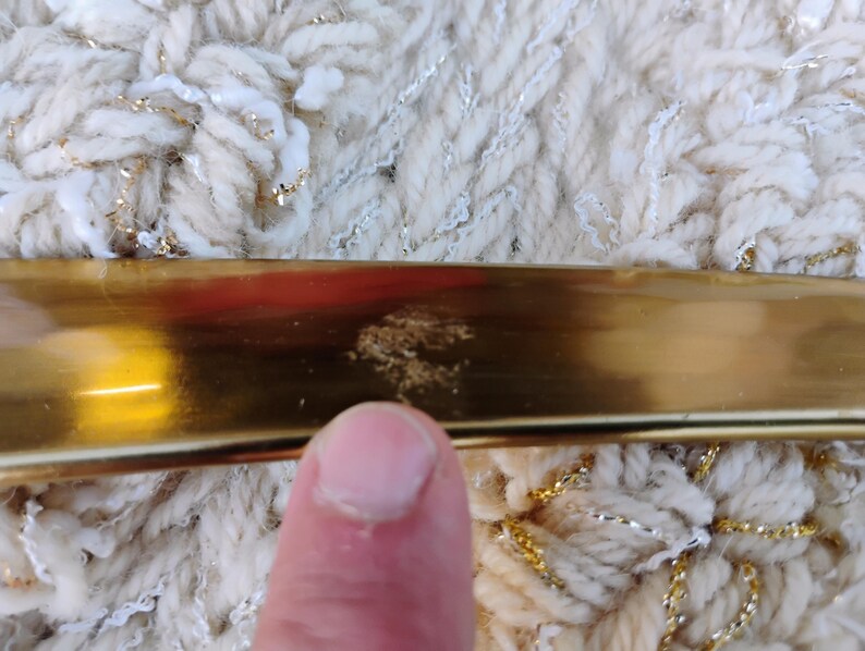 May include: A close-up of a gold-coloured metal object with a reflective surface. The object is positioned against a textured, off-white fabric with gold and silver threads woven throughout. A finger is visible in the foreground.