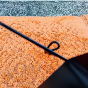 May include: Close-up of an orange shag rug with a floral pattern. A black object with a loop is in the foreground. The rug has a fringe on one side and is on a gray surface.