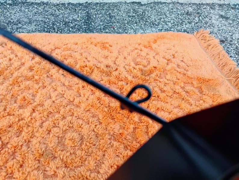 May include: Close-up of an orange shag rug with a floral pattern. A black object with a loop is in the foreground. The rug has a fringe on one side and is on a gray surface.