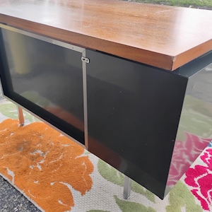 May include: A rectangular desk featuring a smooth, light brown wooden top surface. The base is constructed from solid black panels with a metallic silver vertical trim piece. The unit is supported by thin, straight, silver-coloured metal legs.