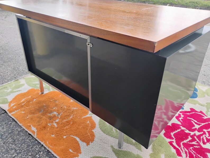 May include: A rectangular desk featuring a smooth, light brown wooden top surface. The base is constructed from solid black panels with a metallic silver vertical trim piece. The unit is supported by thin, straight, silver-coloured metal legs.