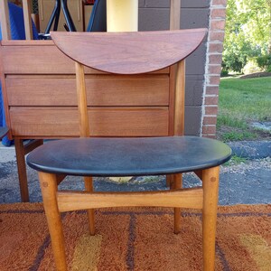 May include: A mid-century modern chair with a dark seat and a curved wooden backrest. The chair has light-colored wooden legs and a dark seat. The chair is placed on a brown rug, with a wooden cabinet in the background.