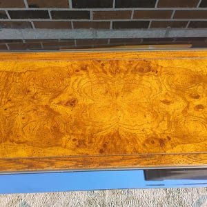 K&ouml;nnte beinhalten: Nahaufnahme einer Holzoberfl&auml;che in warmem, goldbraunem Farbton. Die Holzmaserung weist komplizierte Muster und Wirbel auf, die ein nat&uuml;rliches, strukturiertes Aussehen erzeugen. Die Oberfl&auml;che wird von einem d&uuml;nnen Metallrahmen eingefasst.