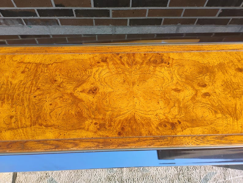 K&ouml;nnte beinhalten: Nahaufnahme einer Holzoberfl&auml;che in warmem, goldbraunem Farbton. Die Holzmaserung weist komplizierte Muster und Wirbel auf, die ein nat&uuml;rliches, strukturiertes Aussehen erzeugen. Die Oberfl&auml;che wird von einem d&uuml;nnen Metallrahmen eingefasst.