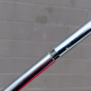 K&ouml;nnte beinhalten: Nahaufnahme einer Chrommetallstange mit sichtbarer Verbindung. Die Stange ist hochreflektierend, mit polierter Oberfl&auml;che. Der Hintergrund ist eine graue Backsteinmauer.