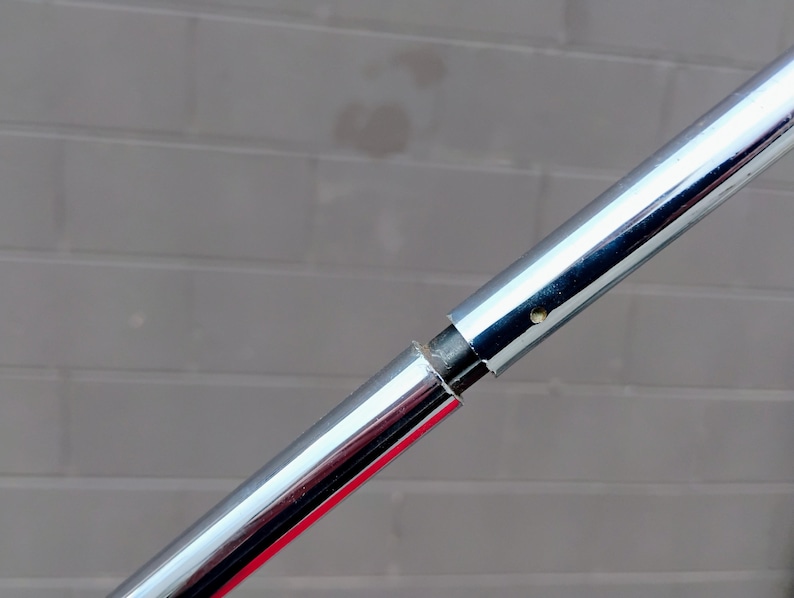 K&ouml;nnte beinhalten: Nahaufnahme einer Chrommetallstange mit sichtbarer Verbindung. Die Stange ist hochreflektierend, mit polierter Oberfl&auml;che. Der Hintergrund ist eine graue Backsteinmauer.