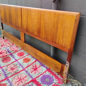 May include: A rectangular wooden headboard featuring a warm, medium-brown wood grain finish. The headboard is constructed with two large, smooth upper panels and a horizontal lower support rail, connected by vertical side posts with metal hardware.