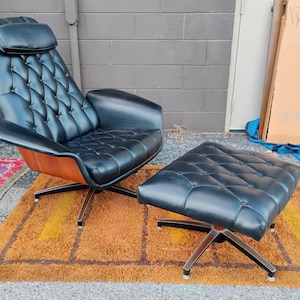 May include: A black leather lounge chair and matching ottoman set featuring a tufted backrest, a headrest pillow, and curved wooden side panels. Both pieces are supported by a four-pronged metal base with a polished finish and black accents.