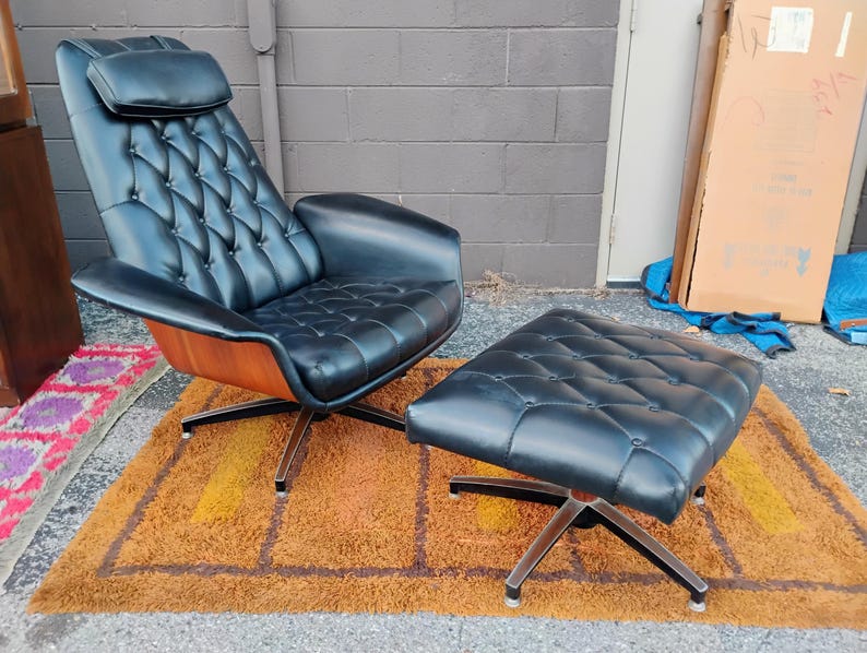 May include: A black leather lounge chair and matching ottoman set featuring a tufted backrest, a headrest pillow, and curved wooden side panels. Both pieces are supported by a four-pronged metal base with a polished finish and black accents.