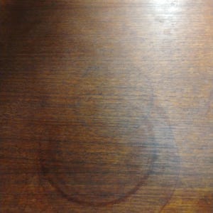 May include: A close-up of a dark brown wooden surface with a large, circular, light brown water stain.