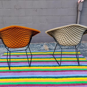 May include: Two wire-frame chairs with black metal grid bases. One chair features a vibrant orange upholstered seat, while the other has a beige upholstered seat. Both chairs have a wide, curved backrest design and thin, angled black metal legs.