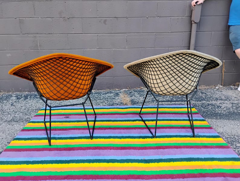 May include: Two wire-frame chairs with black metal grid bases. One chair features a vibrant orange upholstered seat, while the other has a beige upholstered seat. Both chairs have a wide, curved backrest design and thin, angled black metal legs.