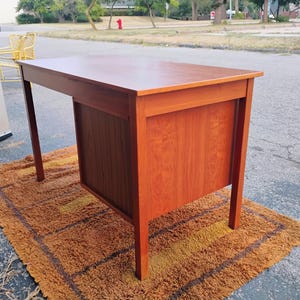 Op de afbeelding: Een rechthoekig houten bureau met een gladde, middenbruine afwerking. Het bureau heeft een vlak bovenblad, vier rechte poten en een zijgedeelte met een kastje met verticale houtnerfpanelen aan de buitenkant voor een stevige constructie.