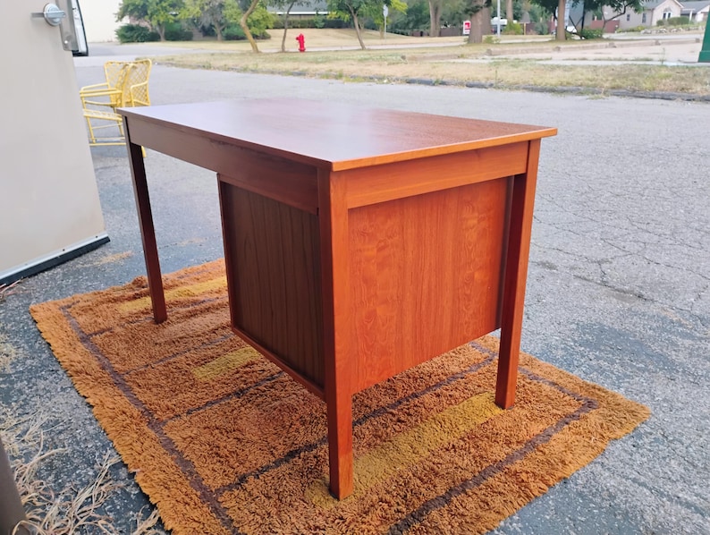Op de afbeelding: Een rechthoekig houten bureau met een gladde, middenbruine afwerking. Het bureau heeft een vlak bovenblad, vier rechte poten en een zijgedeelte met een kastje met verticale houtnerfpanelen aan de buitenkant voor een stevige constructie.