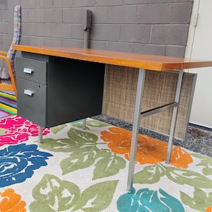 May include: A desk featuring a rectangular wooden top, a dark grey metal cabinet with two drawers, and silver metal legs. The desk includes a textured, light-coloured privacy panel at the back, and the drawers are equipped with silver handles.