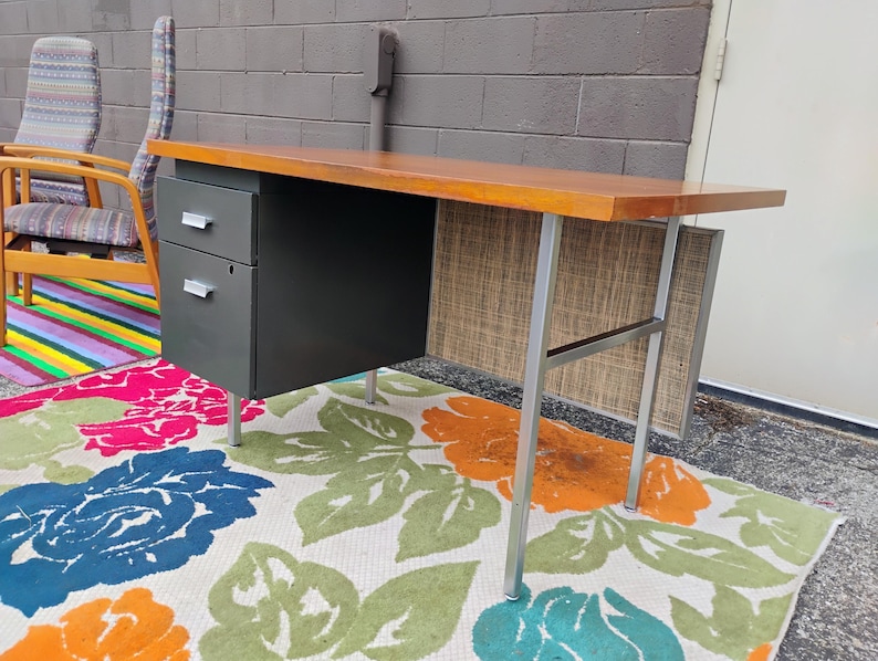 May include: A desk featuring a rectangular wooden top, a dark grey metal cabinet with two drawers, and silver metal legs. The desk includes a textured, light-coloured privacy panel at the back, and the drawers are equipped with silver handles.