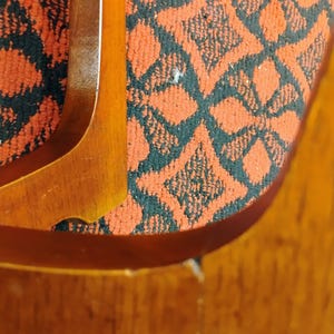 May include: A polished wooden chair frame with curved armrests and legs, featuring a smooth, light brown wood grain finish. The chair is upholstered in a textured fabric with a repeating geometric pattern in black and burnt orange colors.