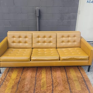 May include: A three-seat sofa upholstered in mustard yellow vinyl featuring a tufted button design on the back cushions and seat cushions. The sofa is supported by a black metal frame with four thin legs, providing a structured and linear appearance.