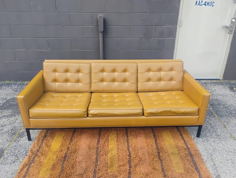 May include: A three-seat sofa upholstered in mustard yellow vinyl featuring a tufted button design on the back cushions and seat cushions. The sofa is supported by a black metal frame with four thin legs, providing a structured and linear appearance.