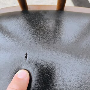 Puede incluir: Un asiento de silla de piel sint&eacute;tica negra con un desgarro visible en el material. El asiento presenta una superficie texturizada y est&aacute; unido a un marco de madera con husillos verticales visibles en el borde superior del respaldo.