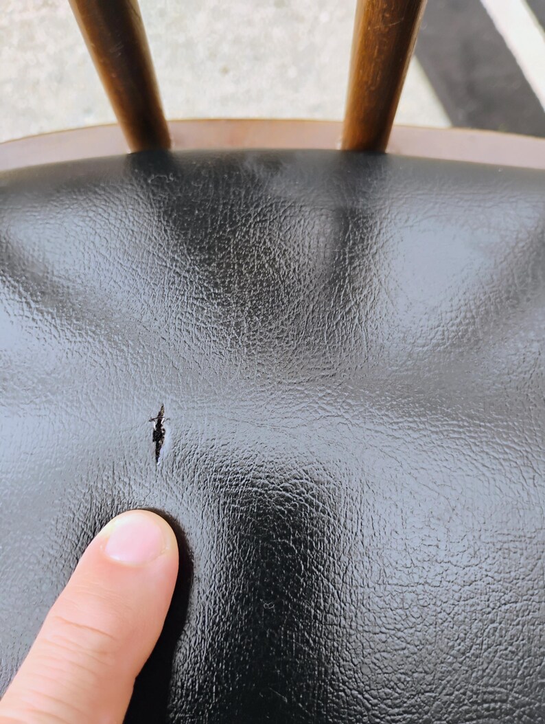 Puede incluir: Un asiento de silla de piel sint&eacute;tica negra con un desgarro visible en el material. El asiento presenta una superficie texturizada y est&aacute; unido a un marco de madera con husillos verticales visibles en el borde superior del respaldo.