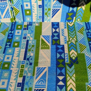 May include: Close-up of a fabric with a geometric pattern in shades of blue, green, and white. The design features vertical stripes with various shapes, including squares, triangles, and chevrons. The fabric has a textured appearance.