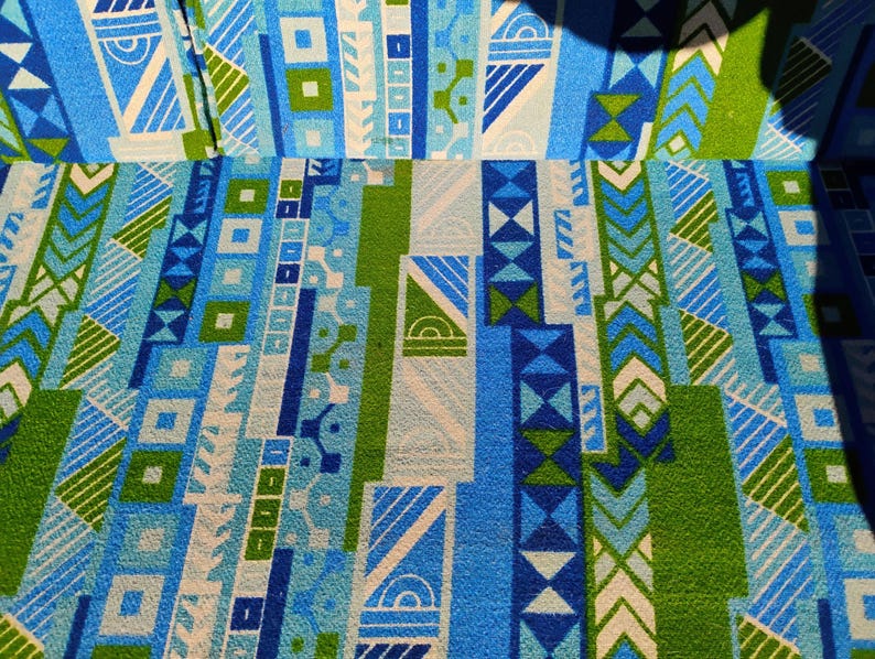 May include: Close-up of a fabric with a geometric pattern in shades of blue, green, and white. The design features vertical stripes with various shapes, including squares, triangles, and chevrons. The fabric has a textured appearance.